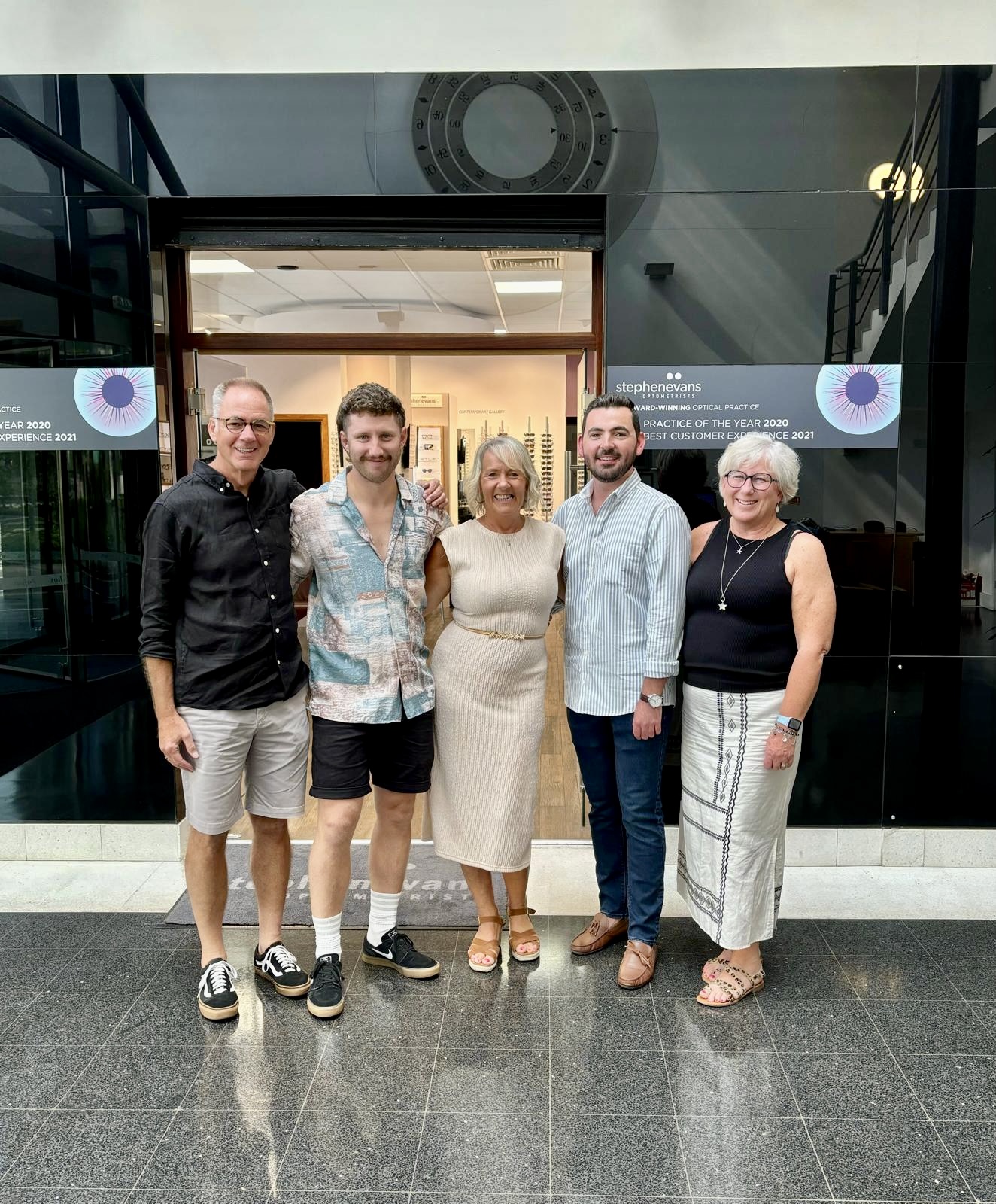 Five members of the Stephen Evans Optometrists team smiling outside their Kings Road practice in Swansea's SA1 Waterfront (left to right): Stephen Evans, Henry Evans, Caroline Paul, Ben Packer and Chrissy Evans, with the practice's award-winning signage visible in the background.