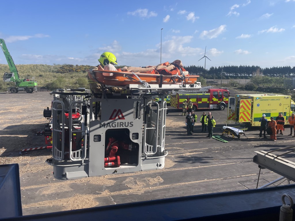 A Magirus turntable ladder appliance with a casualty secured on a stretcher at the top, with a Welsh Ambulance Service vehicle and fire engine visible in the background at Swansea Docks