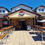 The exterior of The Sandpiper Brewers Fayre pub restaurant on Sandy Road in Llanelli, showing the Brewers Fayre branded frontage with an outdoor beer garden with wooden picnic tables and parasols on a sunny day