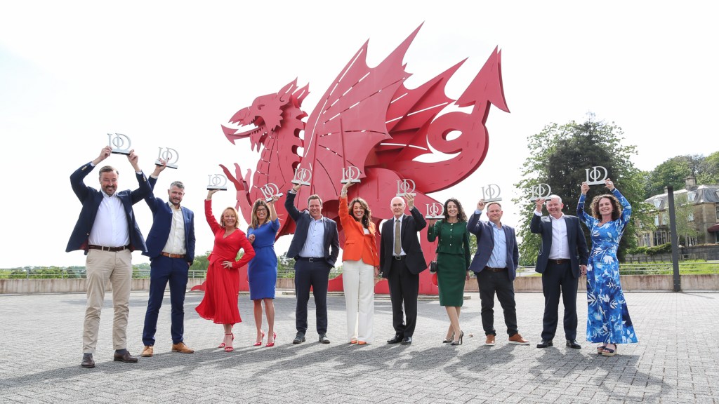 A group of eleven smartly dressed people stand in a line outside ICC Wales in Newport, each holding a silver IoD award trophy aloft, with a large red Welsh dragon sculpture behind them and trees in the background