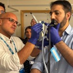 Dr Robin Ghosal wearing blue latex gloves and an NHS lanyard guides a colleague through the CryoEBUS procedure during a training session, with other medical professionals watching in the background
