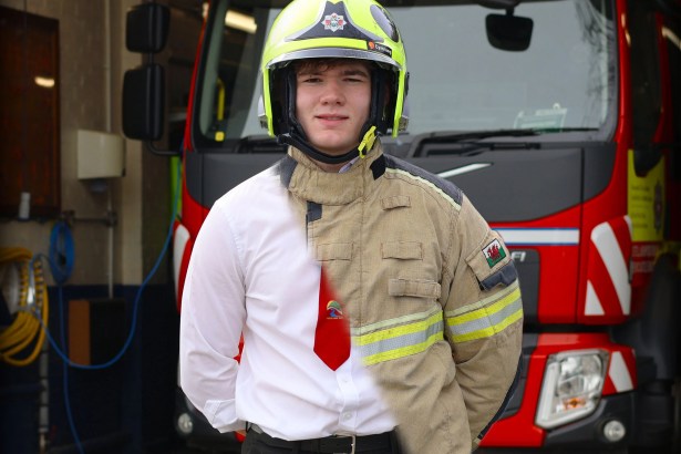 Bobi Jones, an 18-year-old on-call firefighter, stands in front of a red fire engine wearing a yellow firefighter helmet and a beige fire-resistant tunic over his white school shirt and red school tie, with his Ysgol Bro Teifi school uniform visible beneath the tunic