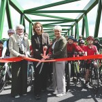 Cllr Linda Evans, Anna McMorrin MP and another official cut a red ribbon on a green metal bridge to mark the official opening of the Tywi Valley Path, with a group of children on bicycles wearing helmets gathered behind them.
