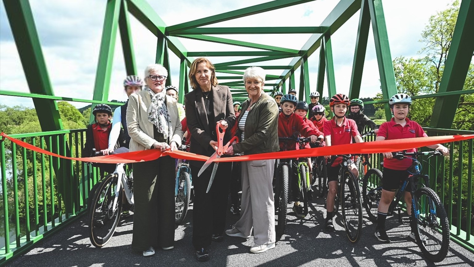 Cllr Linda Evans, Anna McMorrin MP and another official cut a red ribbon on a green metal bridge to mark the official opening of the Tywi Valley Path, with a group of children on bicycles wearing helmets gathered behind them.