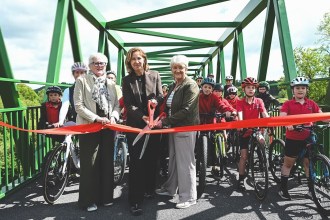 Cllr Linda Evans, Anna McMorrin MP and another official cut a red ribbon on a green metal bridge to mark the official opening of the Tywi Valley Path, with a group of children on bicycles wearing helmets gathered behind them.