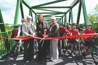 Cllr Linda Evans, Anna McMorrin MP and another official cut a red ribbon on a green metal bridge to mark the official opening of the Tywi Valley Path, with a group of children on bicycles wearing helmets gathered behind them.