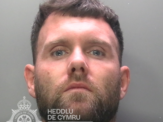 Police custody photograph of Chad White, a white man in his thirties with short dark hair and a beard, wearing a dark green top, against a grey background with a South Wales Police watermark.