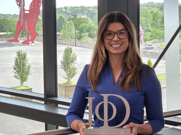 Rachael Flanagan, a woman with long brown hair wearing a blue blazer, holding a silver IoD award trophy and smiling, pictured inside ICC Wales with the large red Welsh dragon sculpture visible through the window behind her