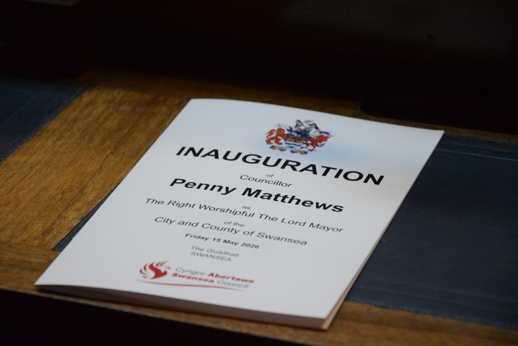 A white inauguration booklet lying on a wooden surface, bearing the Swansea coat of arms and the text "Inauguration of Councillor Penny Matthews as The Right Worshipful The Lord Mayor of the City and County of Swansea, Friday 15 May 2026, The Guildhall, Swansea."