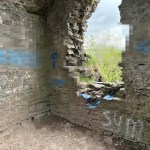 The interior walls of Castell Llansteffan showing blue spray paint graffiti daubed across the ancient stonework, with offensive language pixelated