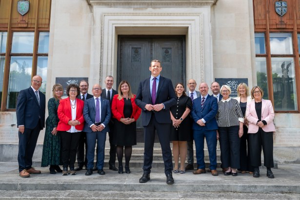 First Minister Rhun ap Iorwerth stands at the centre of a group photograph with his new Cabinet on the steps of the Welsh Government offices in Cardiff, with the Welsh Government dragon logo visible on the building behind them