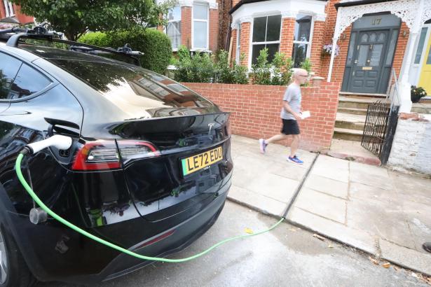 A black Tesla electric vehicle plugged into a green charging cable on a residential street, with a pedestrian walking past on the pavement behind