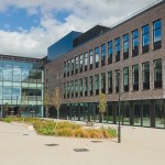 The exterior courtyard of Canolfan Pentre Awel in Llanelli, a modern multi-storey building complex in dark brick with large glazed sections, surrounded by paved walkways and landscaped planting under a blue sky.