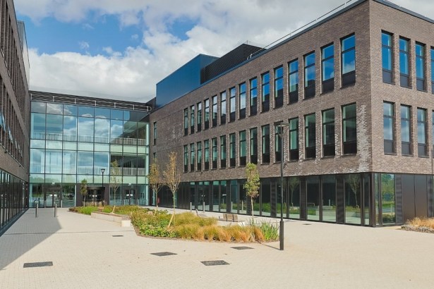 The exterior courtyard of Canolfan Pentre Awel in Llanelli, a modern multi-storey building complex in dark brick with large glazed sections, surrounded by paved walkways and landscaped planting under a blue sky.