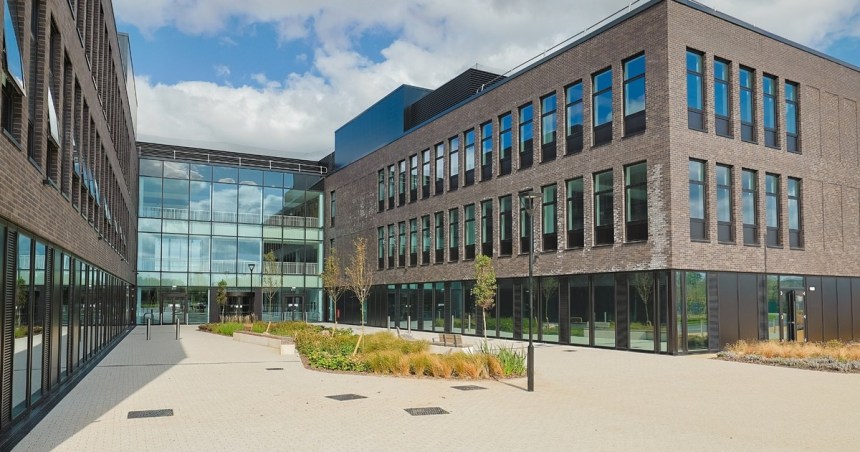 The exterior courtyard of Canolfan Pentre Awel in Llanelli, a modern multi-storey building complex in dark brick with large glazed sections, surrounded by paved walkways and landscaped planting under a blue sky.