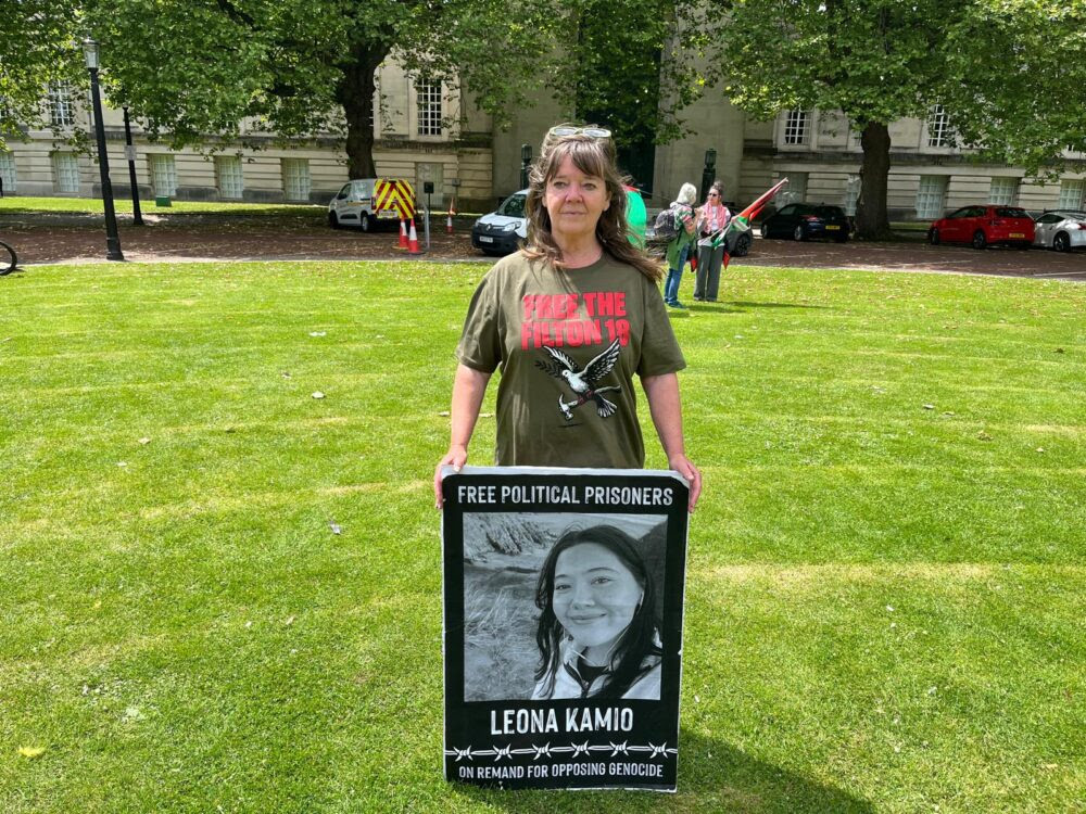 Emma Kamio, wearing a khaki Free the Filton 18 t-shirt, stands on the grass outside the Guildhall in Swansea holding a large placard bearing a black and white photograph of her daughter Leona Kamio with the words Free Political Prisoners and On Remand for Opposing Genocide