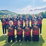 Eleven members of the Western Steel women's cricket team posing for a group photograph on a cricket ground, wearing their new red, black and gold Maiden kit with a hillside and trees visible in the background. Seven players stand in the back row and four kneel in the front row.