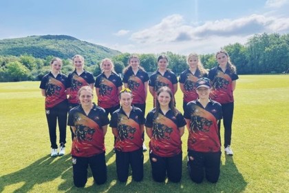 Eleven members of the Western Steel women's cricket team posing for a group photograph on a cricket ground, wearing their new red, black and gold Maiden kit with a hillside and trees visible in the background. Seven players stand in the back row and four kneel in the front row.