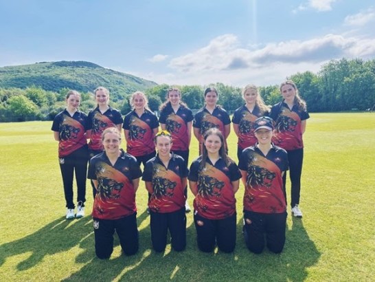 Eleven members of the Western Steel women's cricket team posing for a group photograph on a cricket ground, wearing their new red, black and gold Maiden kit with a hillside and trees visible in the background. Seven players stand in the back row and four kneel in the front row.