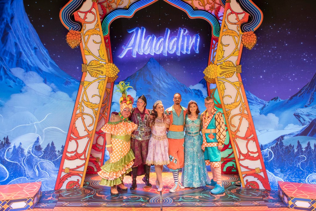 Six pantomime cast members in colourful costumes posed on the Aladdin stage set beneath an illuminated "Aladdin" sign.