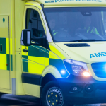 A Welsh Ambulance Service ambulance with blue lights on, moving at speed through a city street with traffic blurred around it.