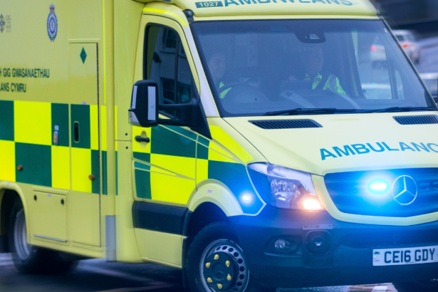 A Welsh Ambulance Service ambulance with blue lights on, moving at speed through a city street with traffic blurred around it.