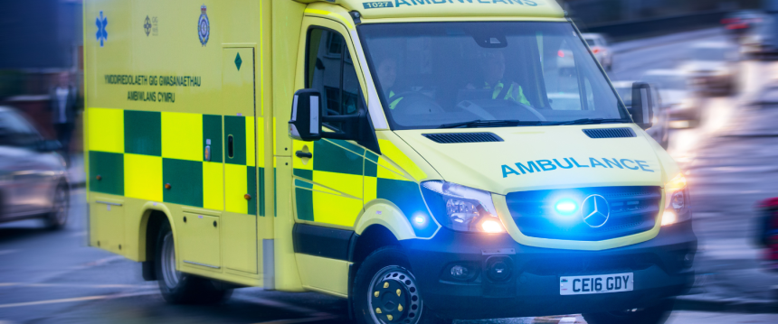 A Welsh Ambulance Service ambulance with blue lights on, moving at speed through a city street with traffic blurred around it.