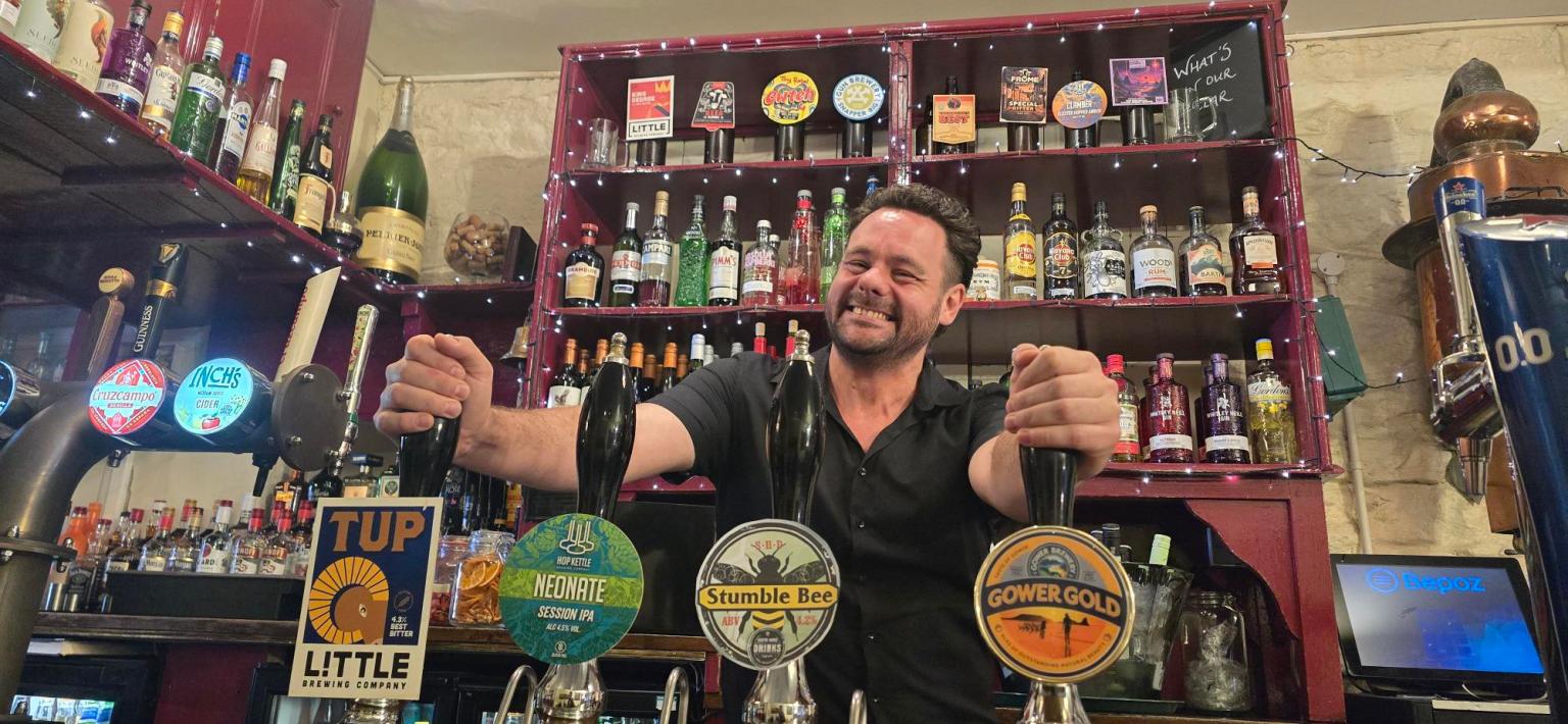 Stuart Davies stands behind the bar at the No Sign Bar on Wind Street, smiling at the camera.