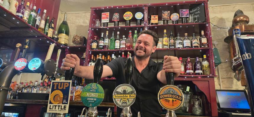 Stuart Davies stands behind the bar at the No Sign Bar on Wind Street, smiling at the camera.