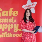 Meghan Smyth smiles holding her award and certificate in front of a red Action for Children banner reading "Safe and happy childhood."