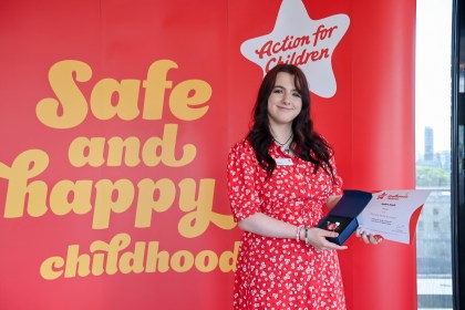 Meghan Smyth smiles holding her award and certificate in front of a red Action for Children banner reading "Safe and happy childhood."