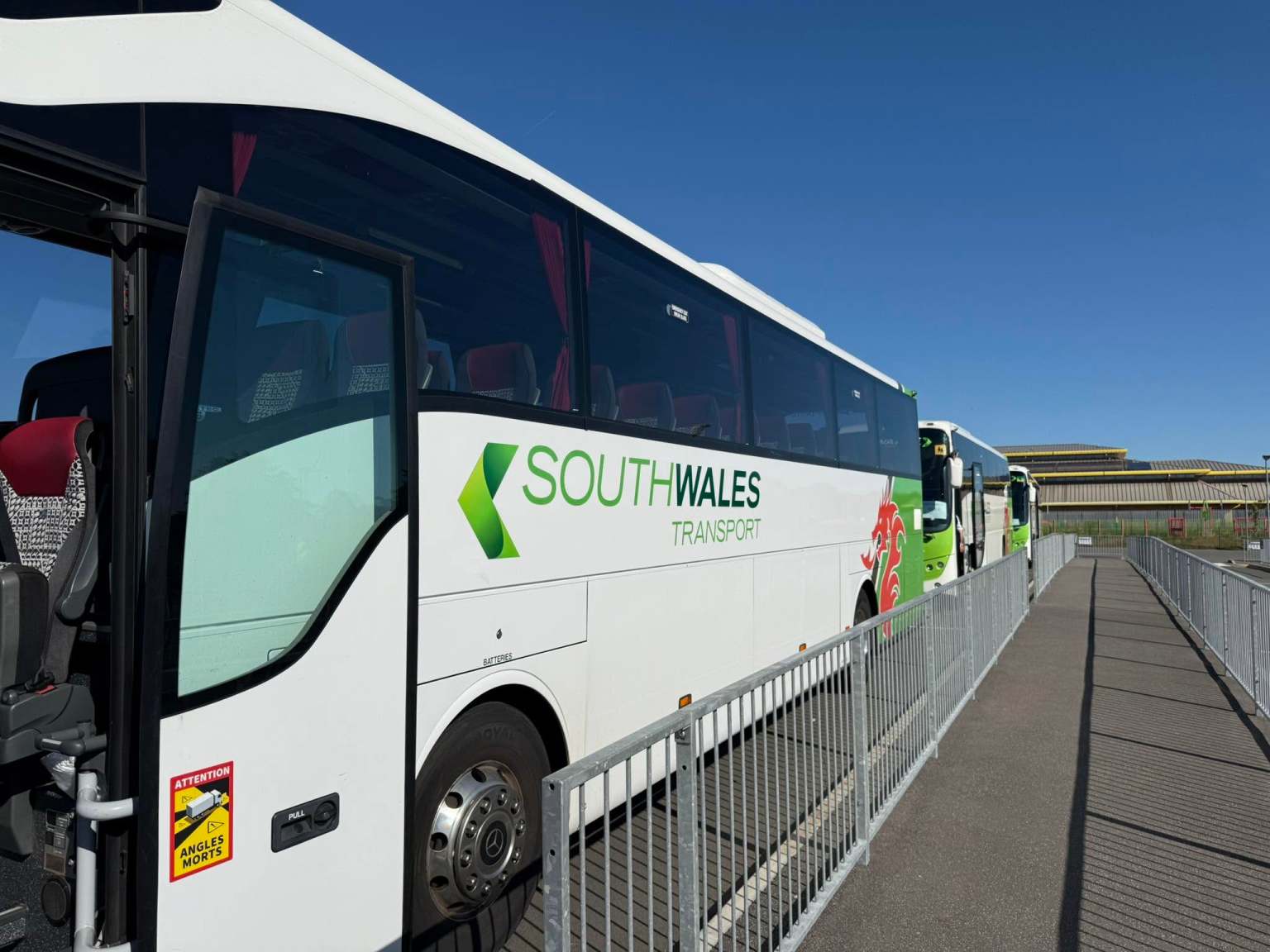 The side of a white South Wales Transport coach with its green logo and red Welsh dragon, parked behind railings under a clear blue sky, with more coaches lined up behind.