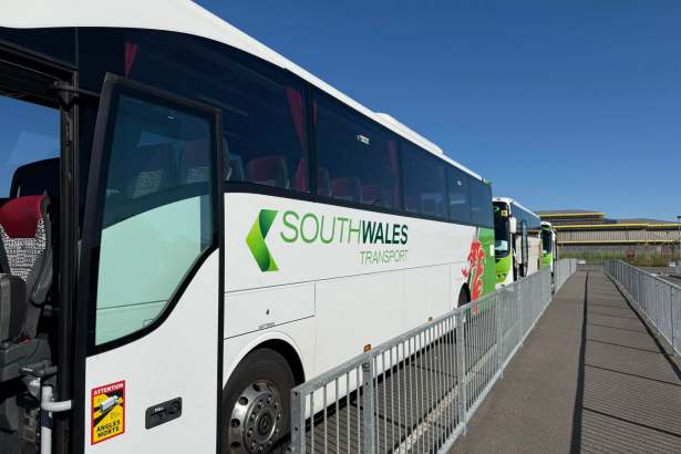 The side of a white South Wales Transport coach with its green logo and red Welsh dragon, parked behind railings under a clear blue sky, with more coaches lined up behind.