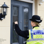 A police officer in a high-visibility jacket and bowler hat knocking on the front door of a house.