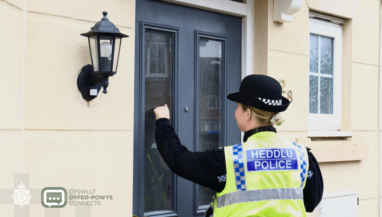 A police officer in a high-visibility jacket and bowler hat knocking on the front door of a house.