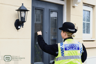 A police officer in a high-visibility jacket and bowler hat knocking on the front door of a house.