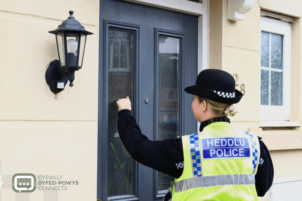 A police officer in a high-visibility jacket and bowler hat knocking on the front door of a house.