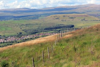 A view across the Vale of Neath from a grassy hillside with a boundary fence, looking down towards a village with hills beyond.