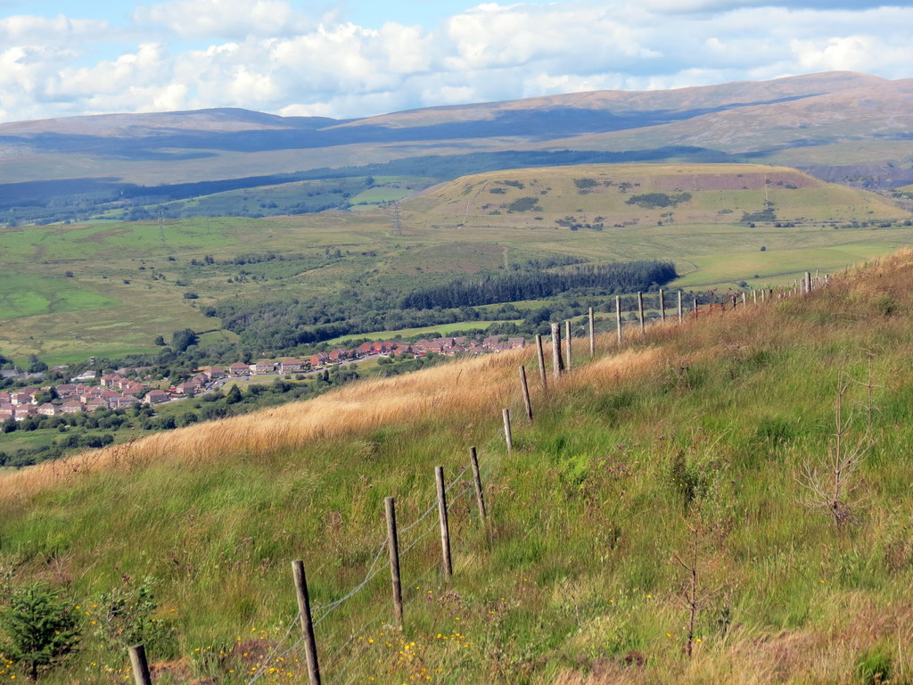 A view across the Vale of Neath from a grassy hillside with a boundary fence, looking down towards a village with hills beyond.