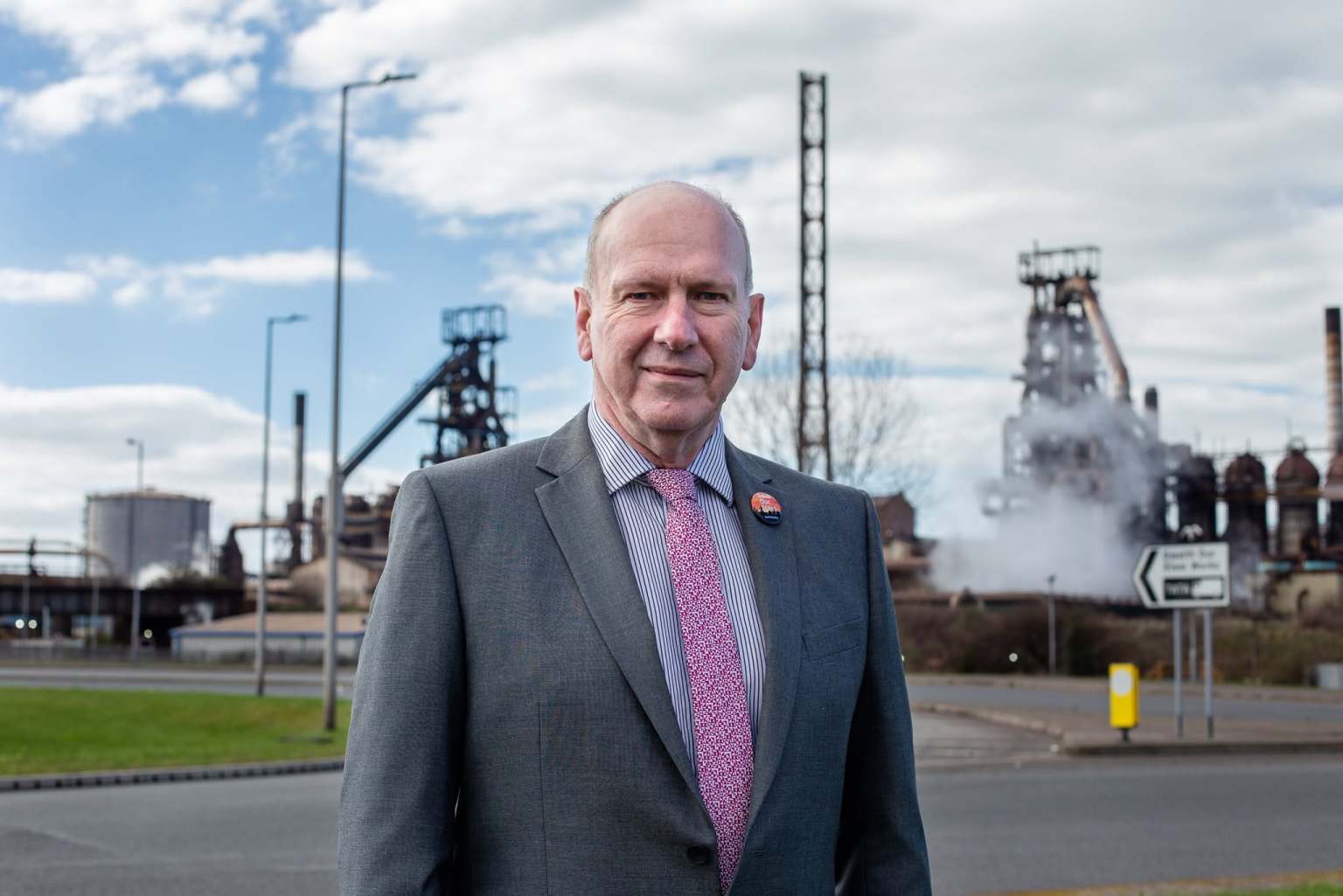 David Rees in a grey suit standing in front of the Port Talbot steelworks, with blast furnaces and a steam plume behind him.