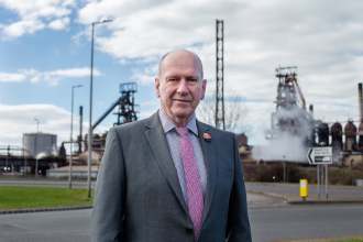 David Rees in a grey suit standing in front of the Port Talbot steelworks, with blast furnaces and a steam plume behind him.