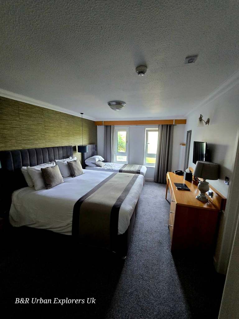 A made-up hotel bed with drinks bottles and a telephone on the desk and an open wardrobe alongside