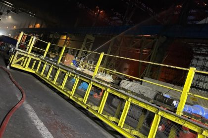 The gutted, glowing interior of a steelworks building at night with a hose directed across a walkway