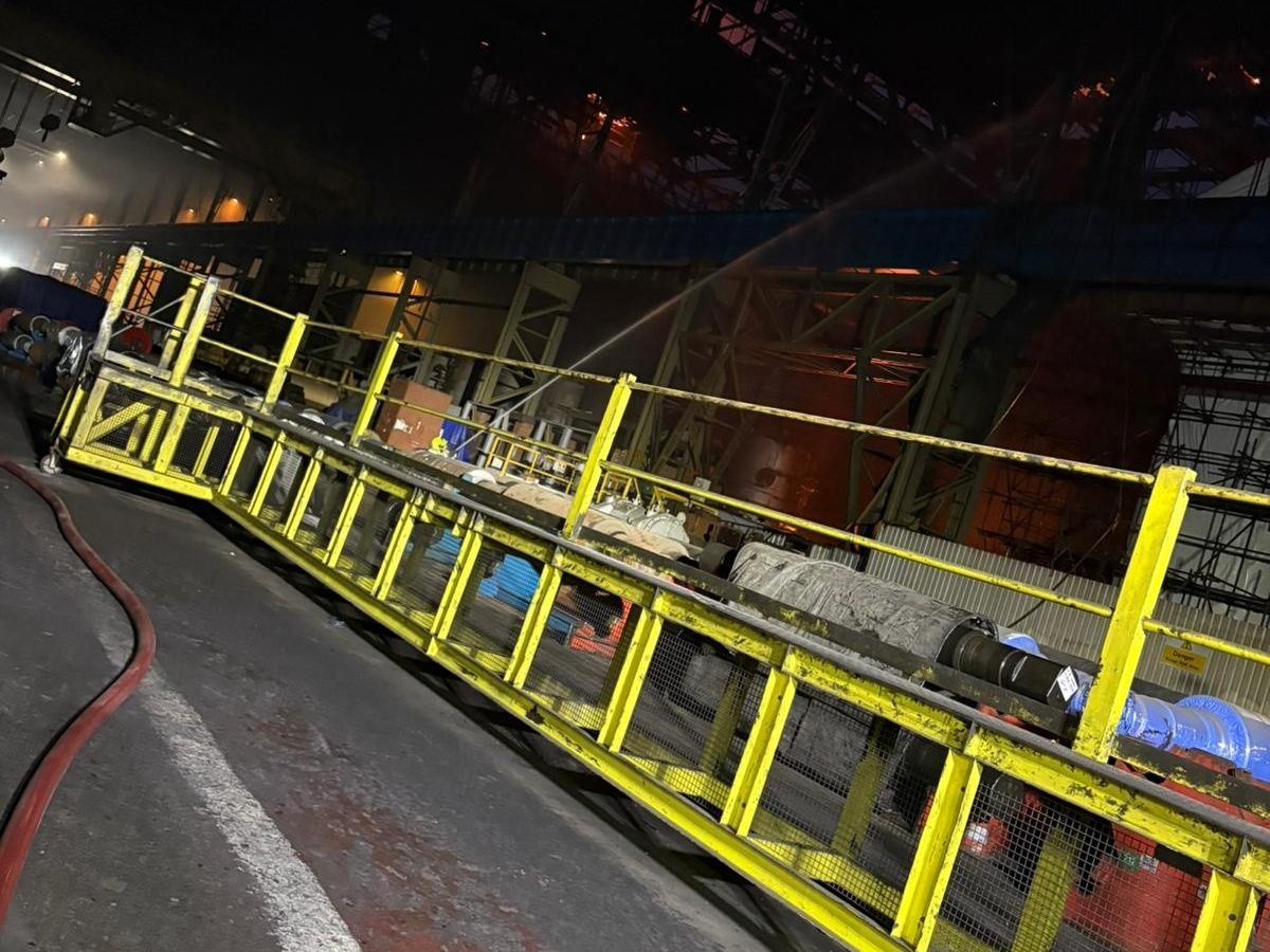 The gutted, glowing interior of a steelworks building at night with a hose directed across a walkway
