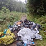 A large pile of black planters, growing medium and sheeting fly-tipped on a forestry track surrounded by trees
