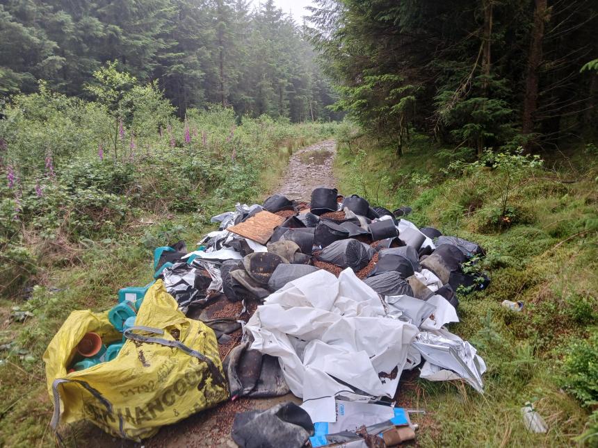 A large pile of black planters, growing medium and sheeting fly-tipped on a forestry track surrounded by trees
