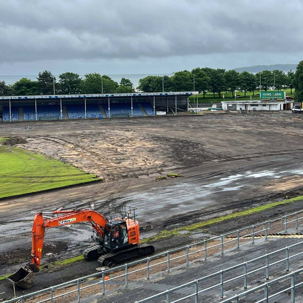 A digger on the bare, muddy pitch at St Helen's in front of the main stand, with the sea in the distance.