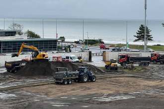 Excavators and tipper trucks working on the dug-up pitch at St Helen's, with the sea behind.