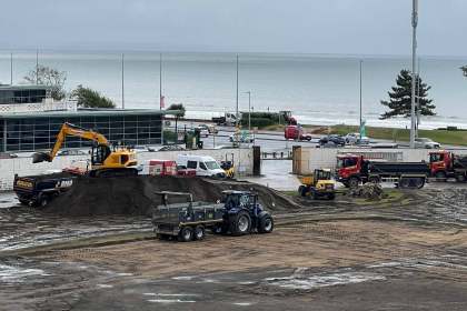 Excavators and tipper trucks working on the dug-up pitch at St Helen's, with the sea behind.