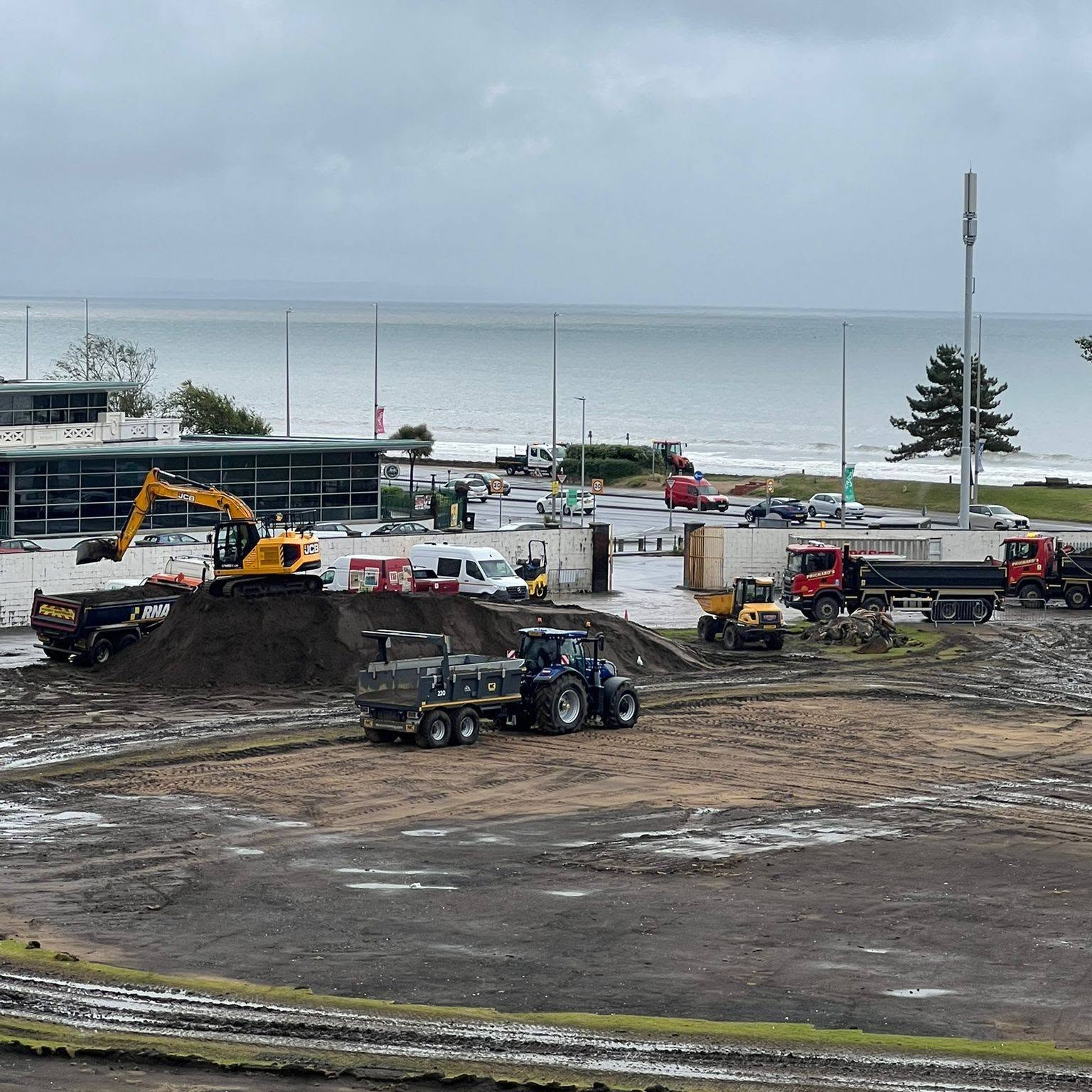 Excavators and tipper trucks working on the dug-up pitch at St Helen's, with the sea behind.